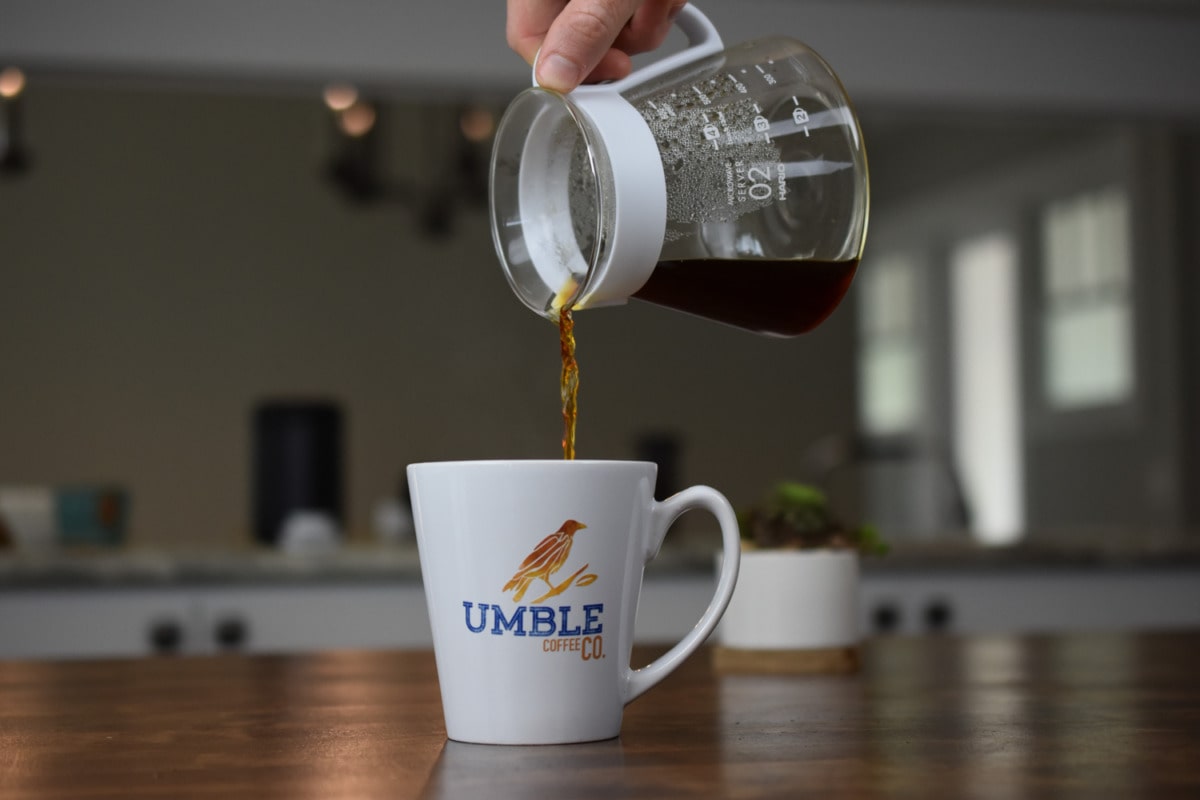 Coffee being poured from a glass carafe into a white mug with 'Umble Coffee' branding on a kitchen counter.