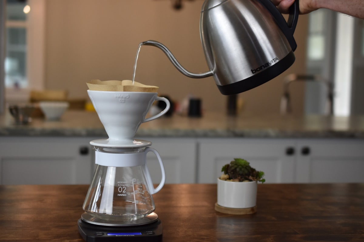 Gooseneck kettle pouring hot water into a Hario V60 dripper on a glass carafe.