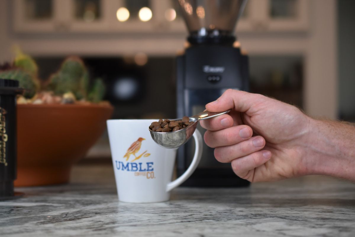Hand holding a spoon with coffee beans over a white mug labeled 'Lumble' on a kitchen counter.