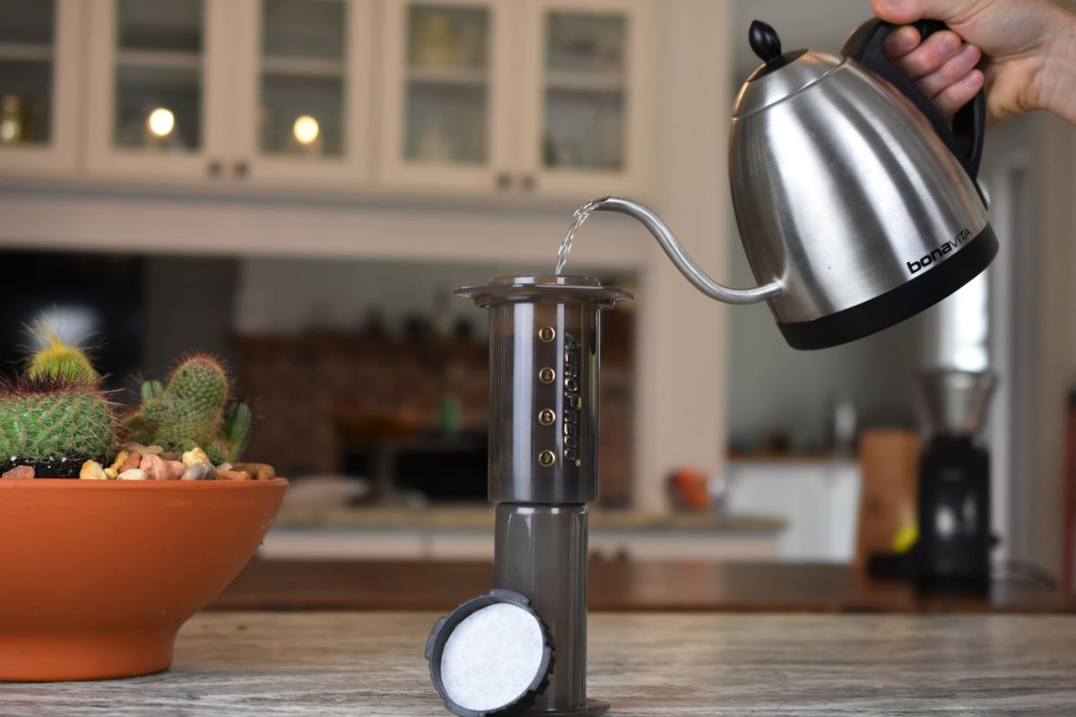 Person pouring water from a kettle into an AeroPress coffee maker on a kitchen counter.