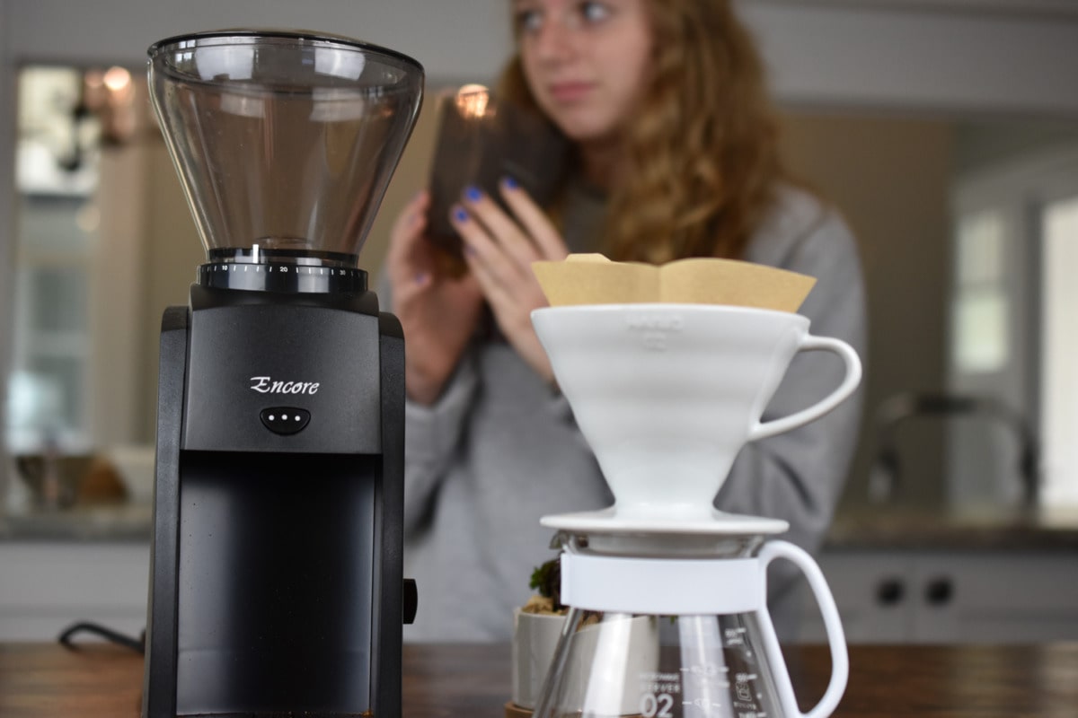 Baratza Encore grinder next to a Hario V60 dripper with filter, with a person holding a coffee bag in the background.
